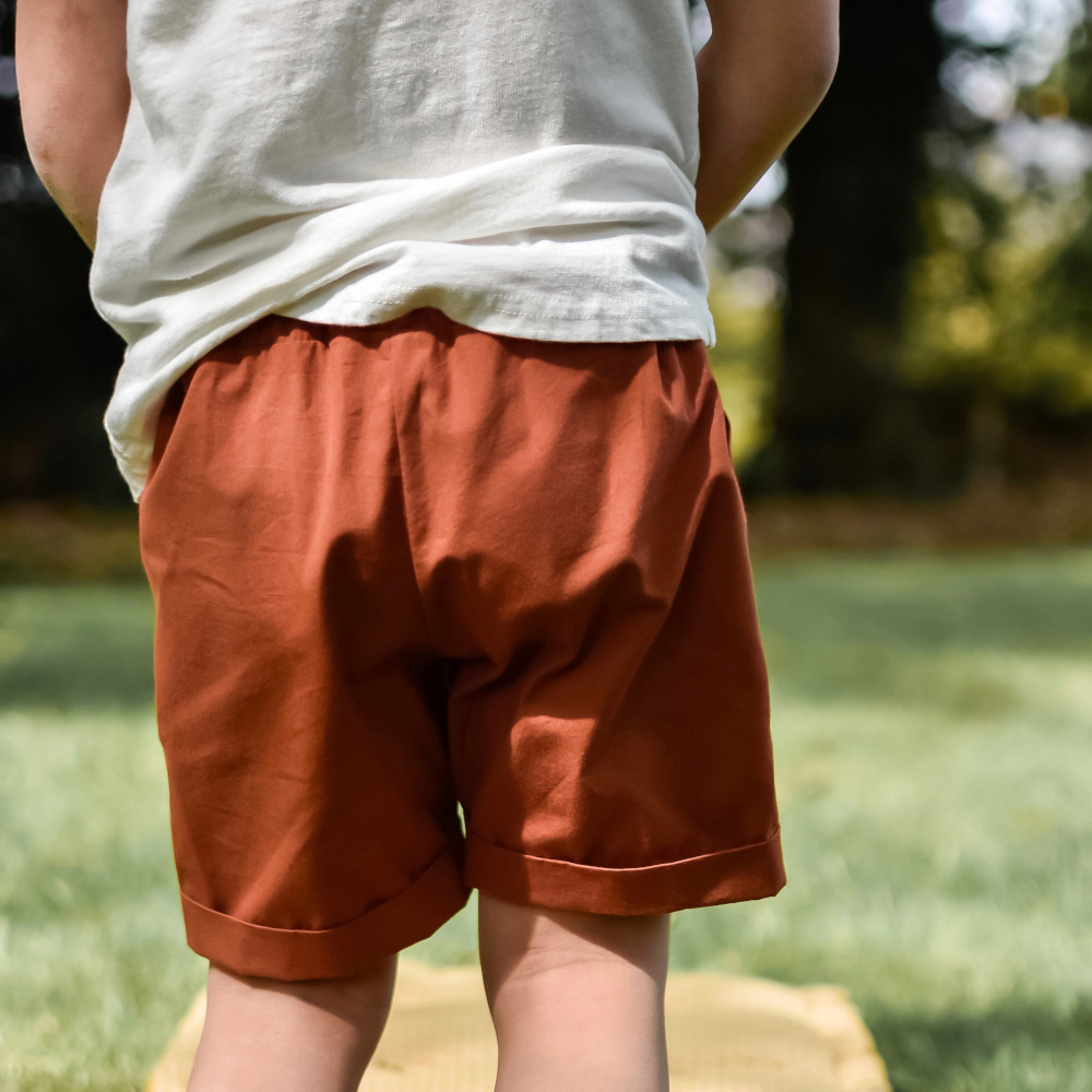 Vue de dos d'un enfant portant un bermuda terracotta avec revers, debout dans l'herbe