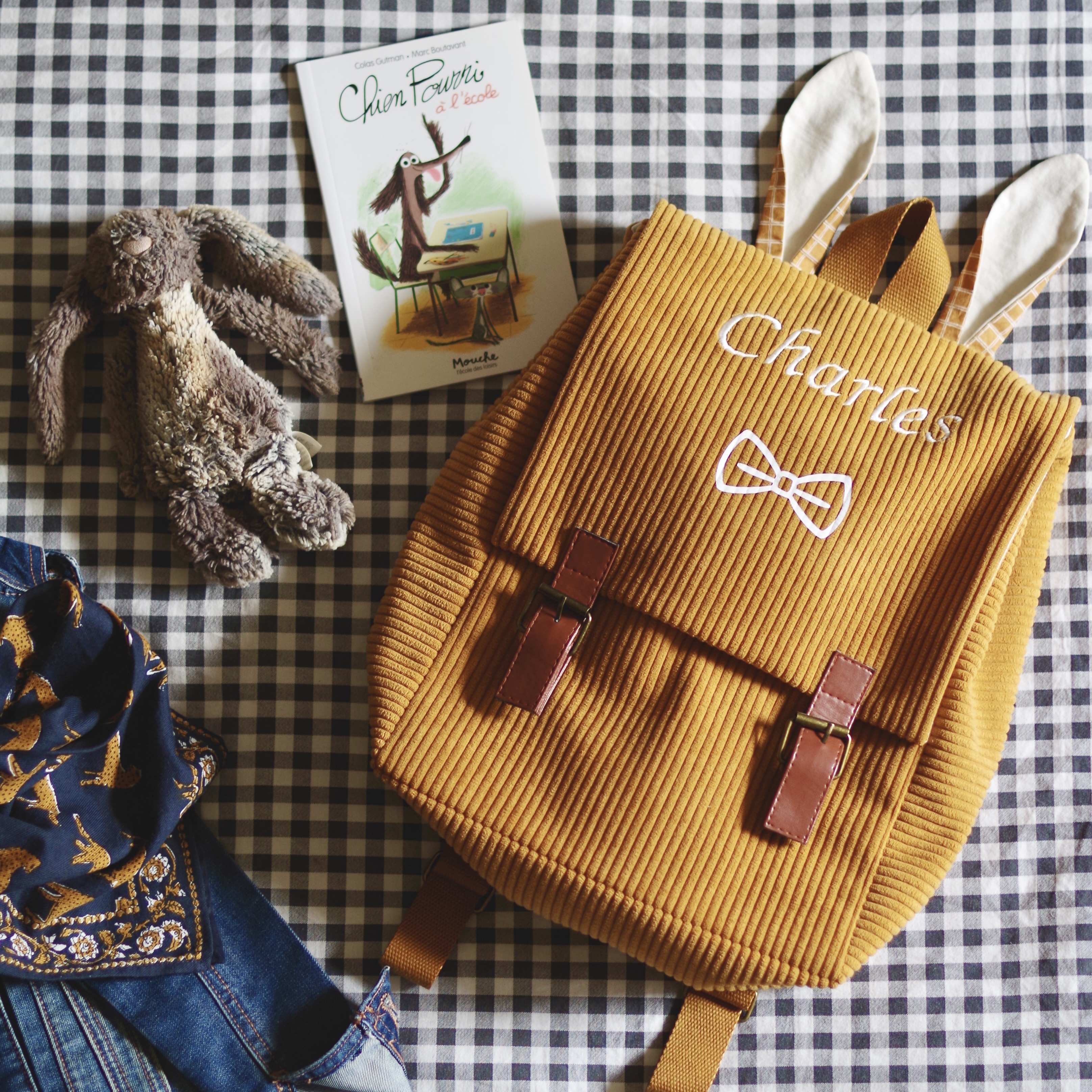 Sac à dos pour la maternelle, en velours grosses côtes camel, présenté sur un lit avec une veste en jean, un doudou et un livre. Il est brodé au prénom Charles.