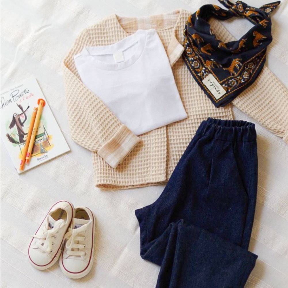 Gilet enfant en tissu nid d'abeille beige à manches longues, présenté à plat avec un t-shirt blanc, un jean brut, un bandana à imprimé animaux, des baskets blanches et un livre illustré pour enfant