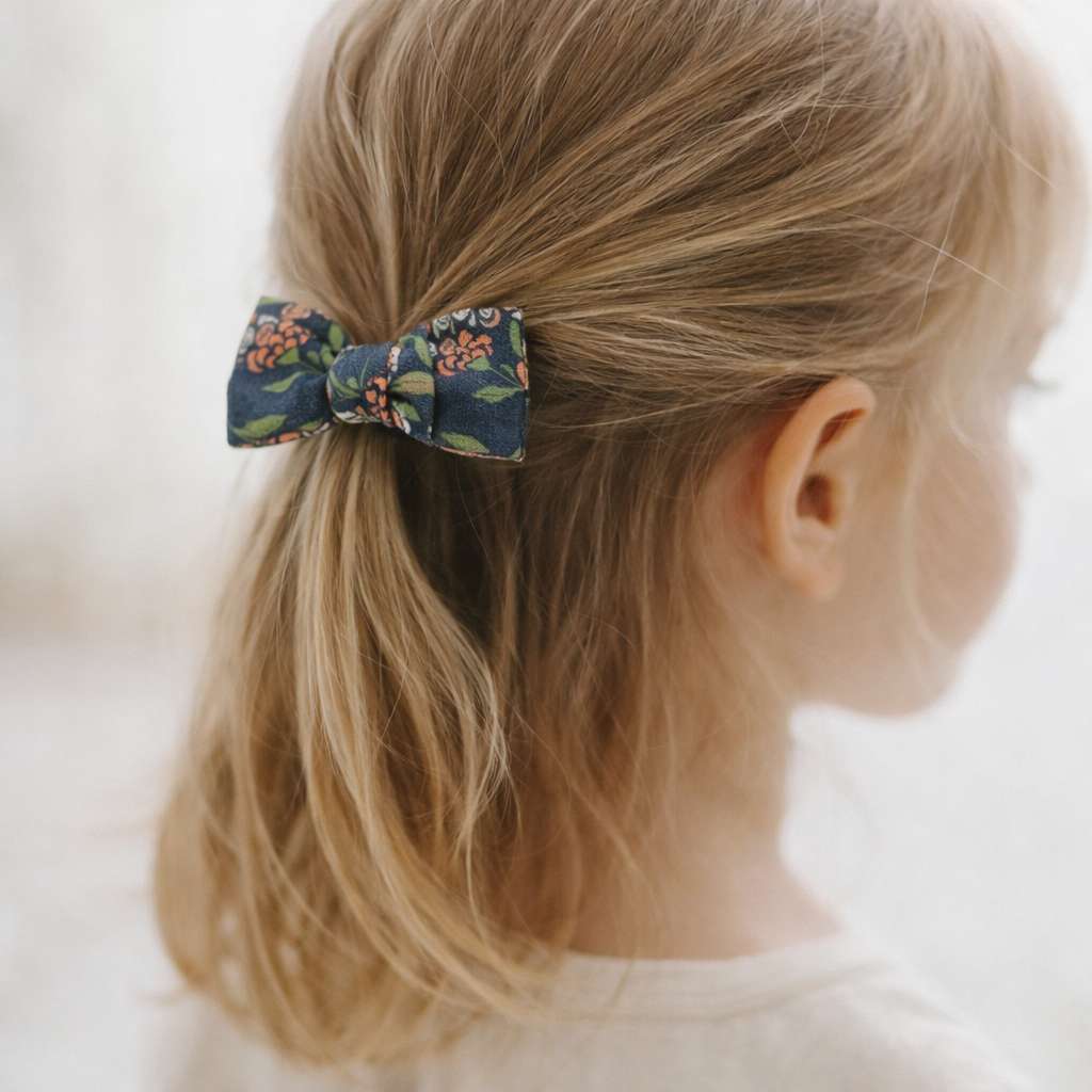 Nœud élastiqué pour cheveux enfant en coton fleuri dans des tons corail et vert olive sur fond anthracite, porté en queue de cheval par une petite fille aux cheveux blonds.