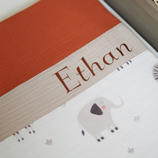 Zoom sur la couverture plaid bébé personnalisée avec le prénom 'Ethan'. Tissus en double gaze de coton imprimé avec des éléphants et lions sur fond blanc.