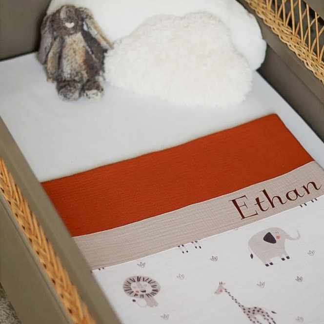 Couverture bébé personnalisée en double gaze de coton terracotta, beige et animaux de la savane installée dans un berceau avec une peluche et un coussin nuage. Idéale pour un cocon douillet