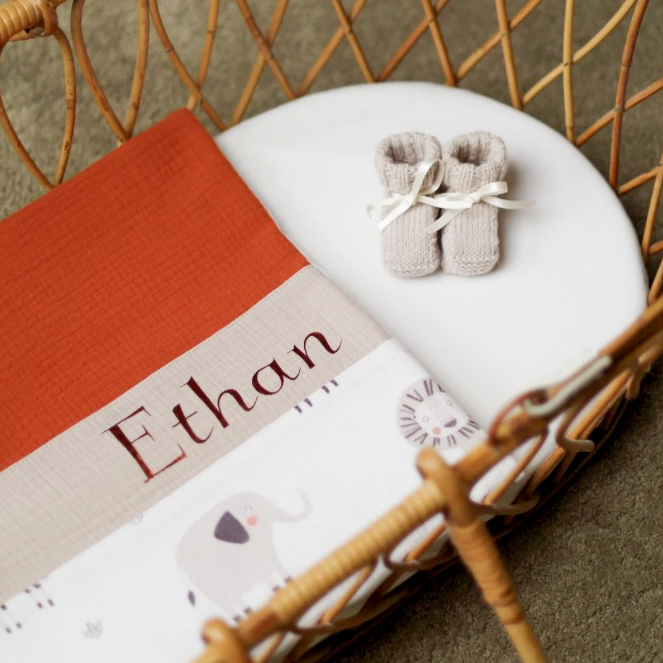 Couverture bébé personnalisée avec le prénom 'Ethan', pliée sur un matelas de couffin en rotin. Motifs animaux et teintes douces en terracotta, beige et blanc.