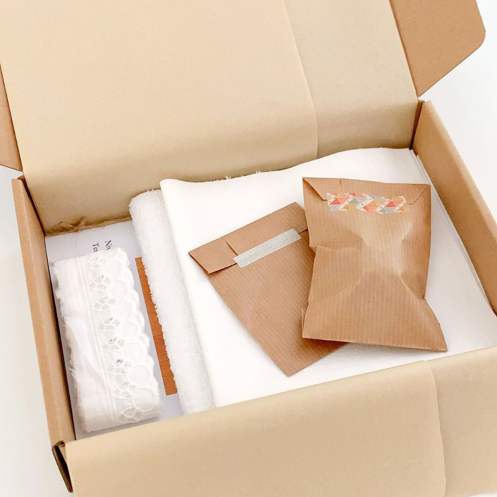 Visuel sur le contenu du kit couture pour réaliser un bavoir comme autrefois : livret détaillé pas à pas, dentelle, bouton, tissus ... emballés dans du papier de soie et une jolie boîte kraft