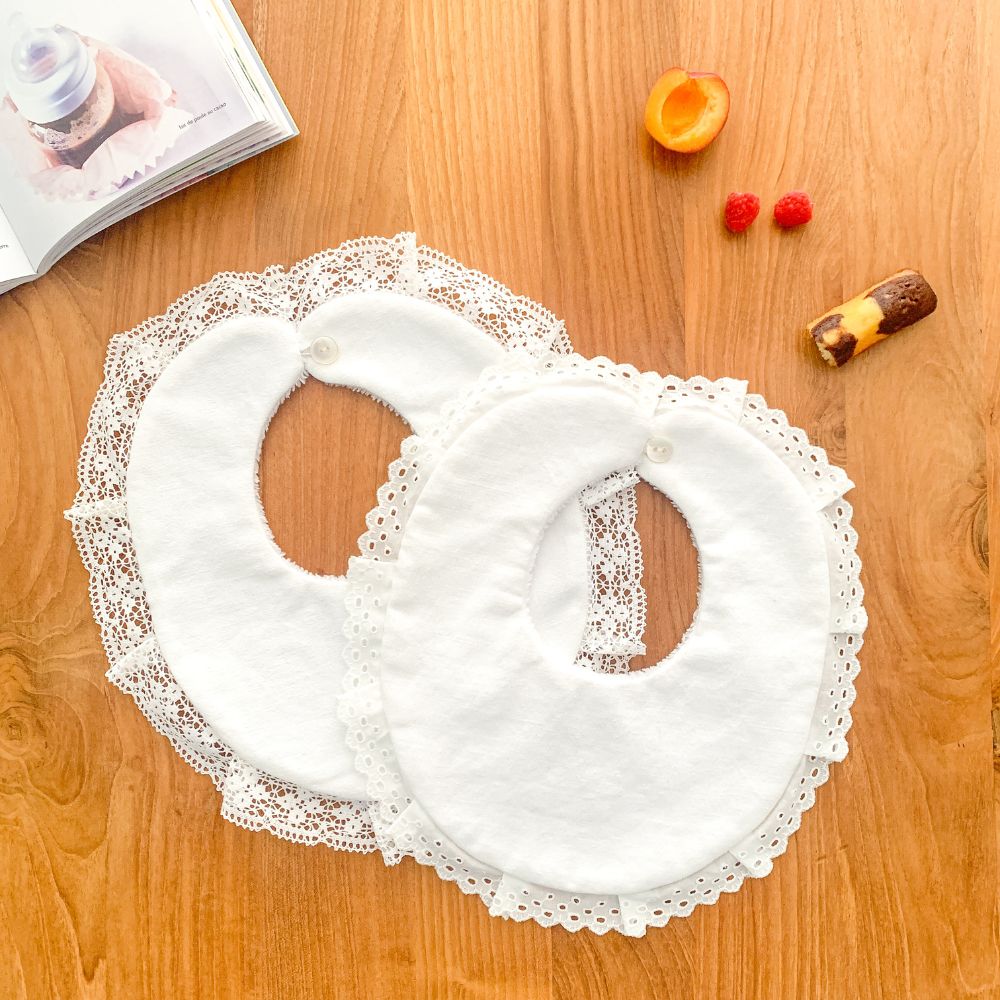 Deux bavoirs blancs finis avec dentelles anciennes chinées, posés sur une table en bois clair, à côté d’un livre, de fruits et d’un petit gâteau. Créations artisanales issues d'un kit créatif Rélinette Créations.