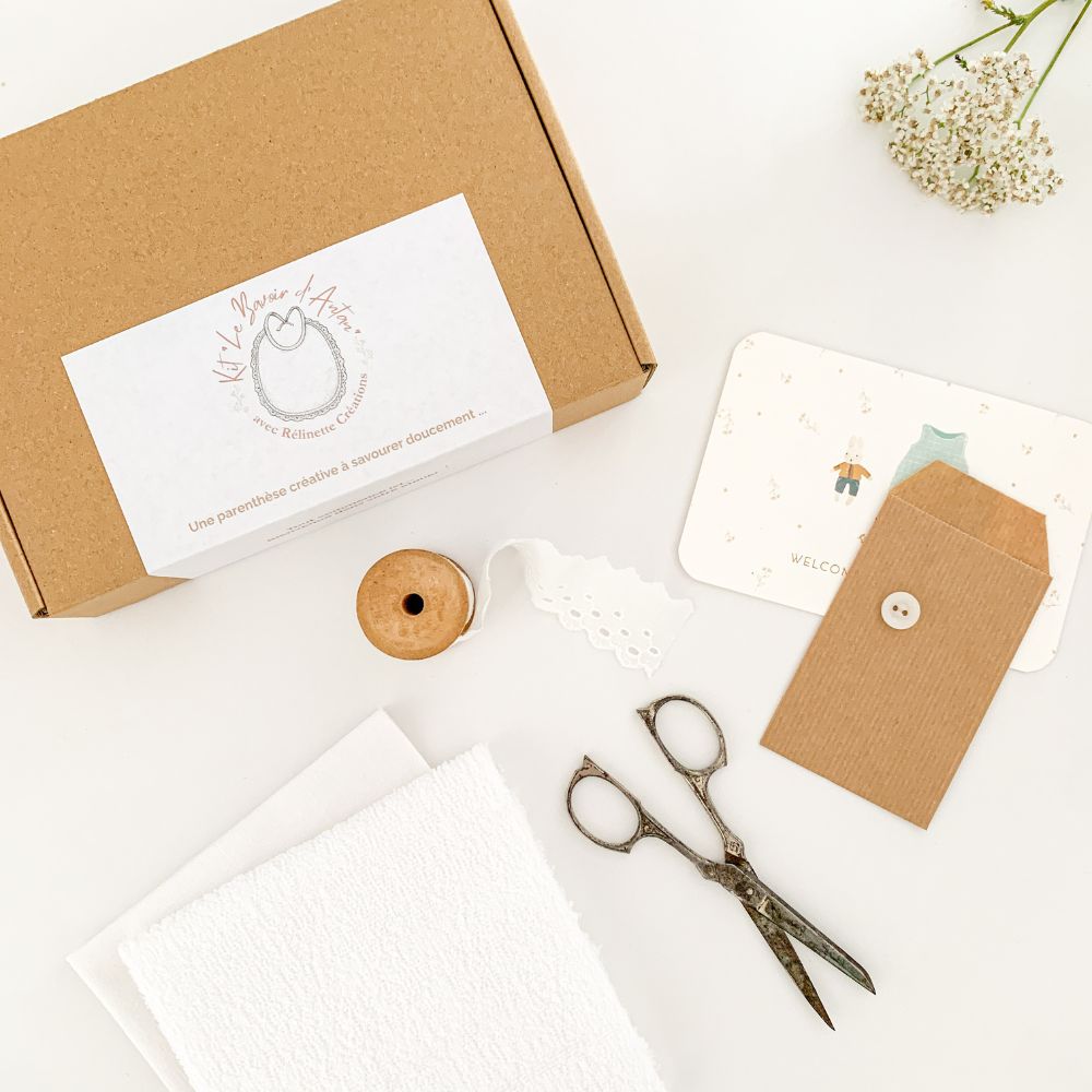 Kit créatif "Le bavoir d'Antan" par Rélinette Créations présenté dans une boîte kraft avec étiquette, dentelle blanche, bouton, carte de bienvenue et paire de ciseaux anciens posés sur un fond blanc.