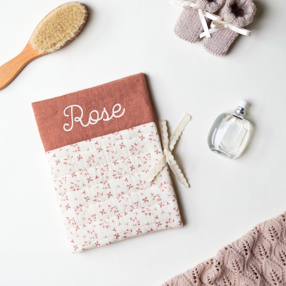 Protège carnet de santé personnalisé avec le prénom Rose, en tissu double gaze à motifs fleurs roses sur fond écru et lin vieux rose, fermé par un ruban de dentelle, photographié avec une brosse à cheveux en bois et des chaussons bébé tricotés
