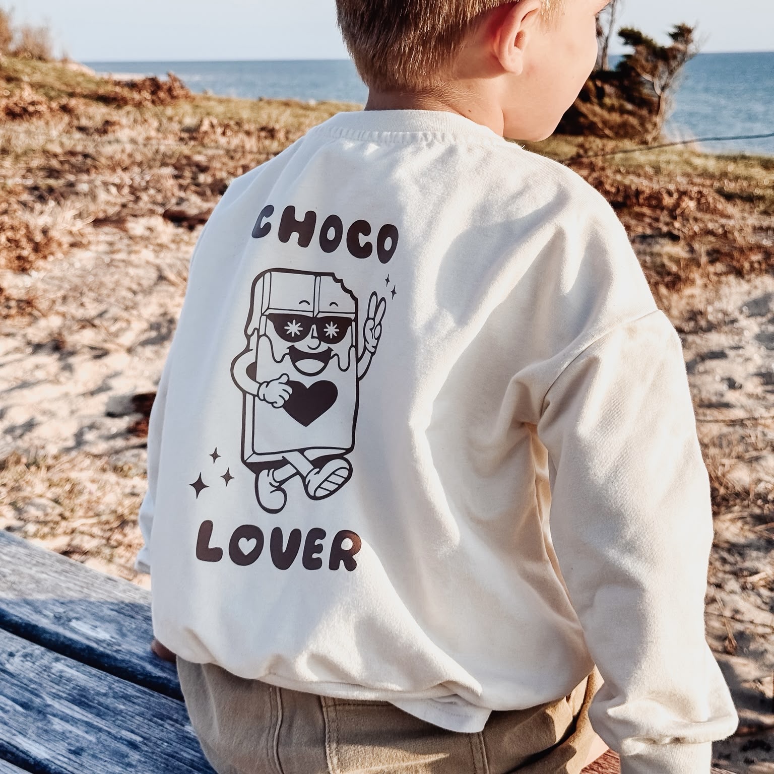 Sweatshirt écru pour enfant avec imprimé "Choco Lover" et illustration d'une tablette de chocolat souriante portant des lunettes de soleil, porté par un garçon face à la mer.