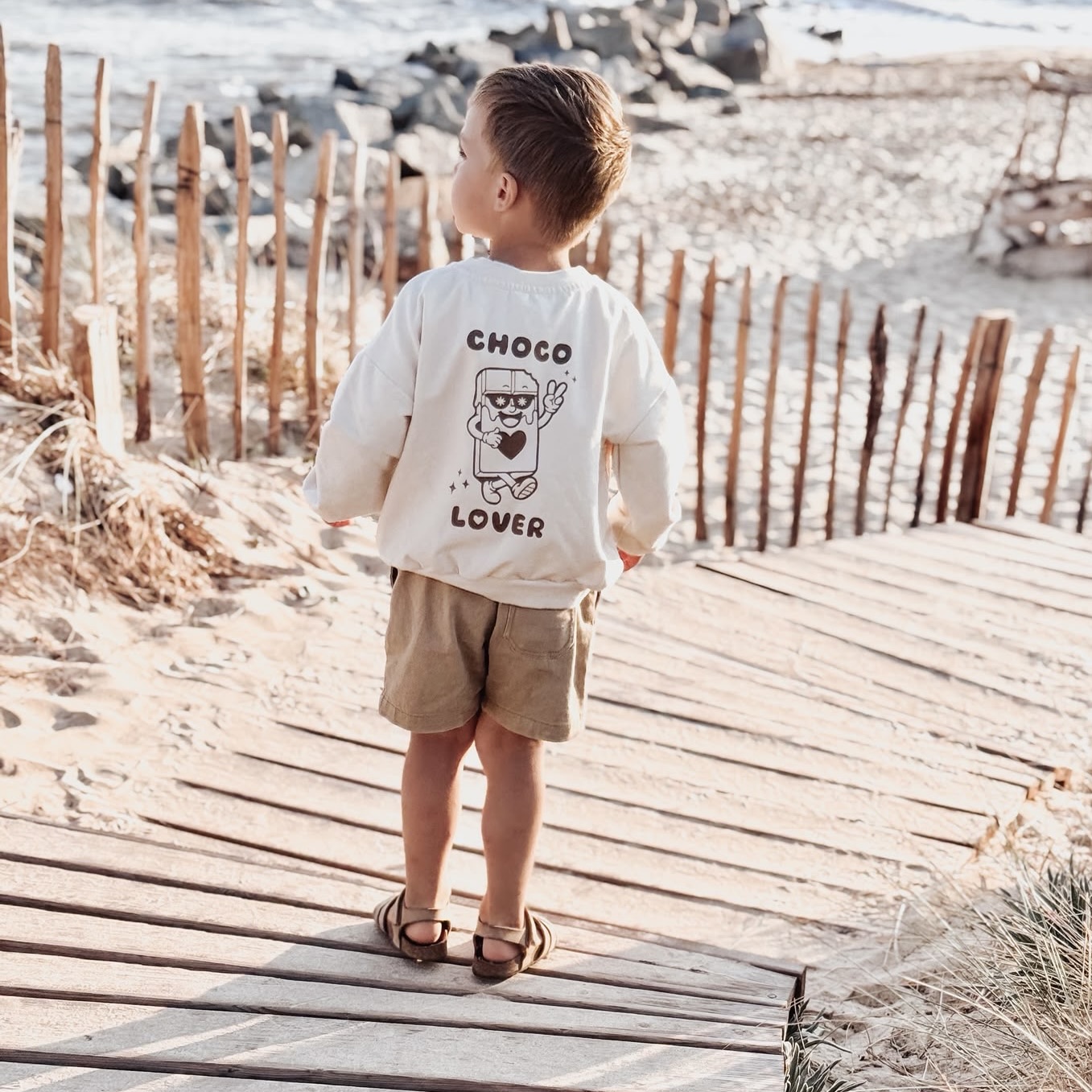 Petit garçon de dos en sweatshirt écru "Choco Lover" et short caramel, marchant pieds en sandales sur un ponton en bois menant à la plage.