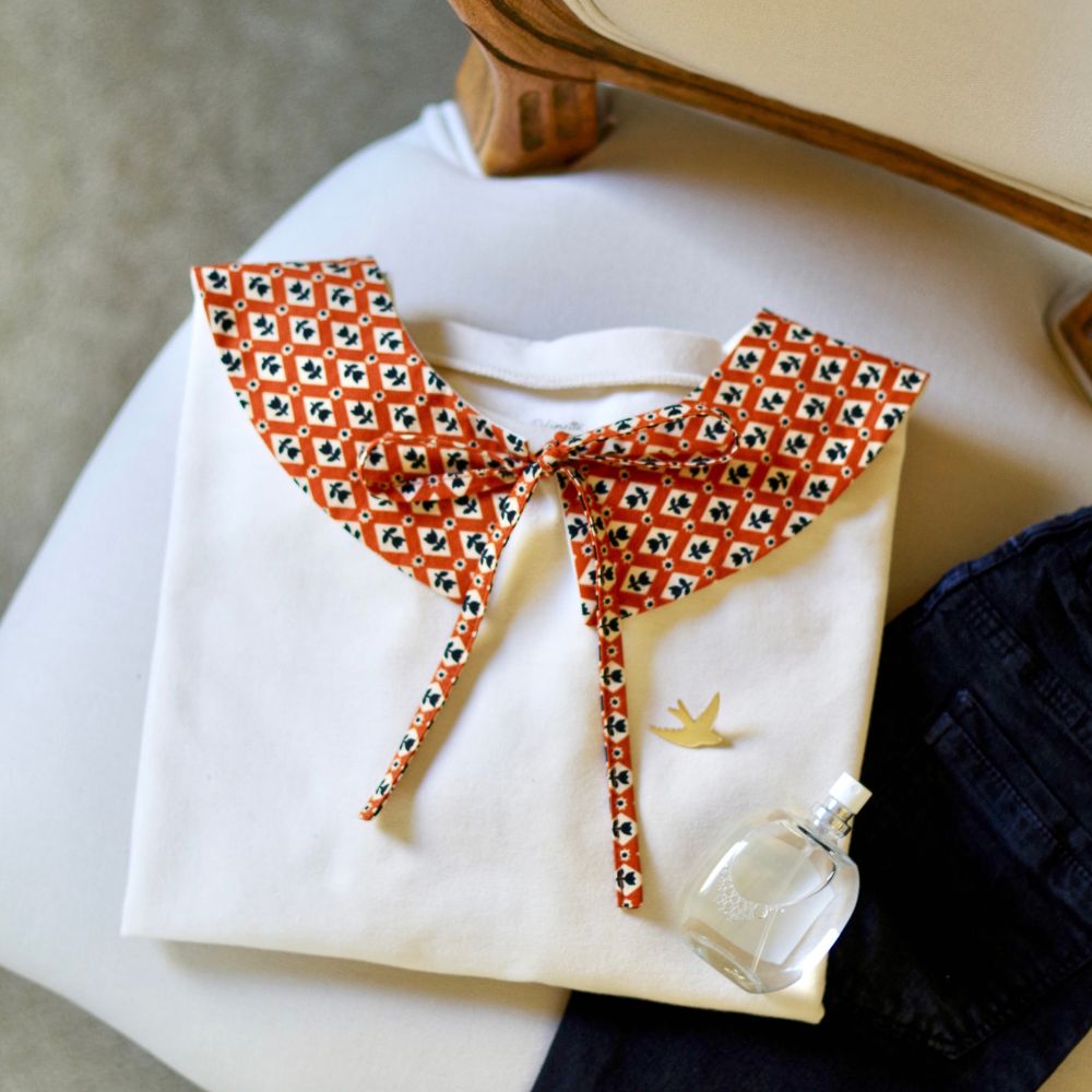 Sweat écru plié sur un fauteuil, orné d’un col Claudine en tissu caramel à motifs géométriques et fleurs noires, noué par un ruban. Une broche hirondelle dorée et un flacon de parfum sont posés à côté, près d’un jean noir.