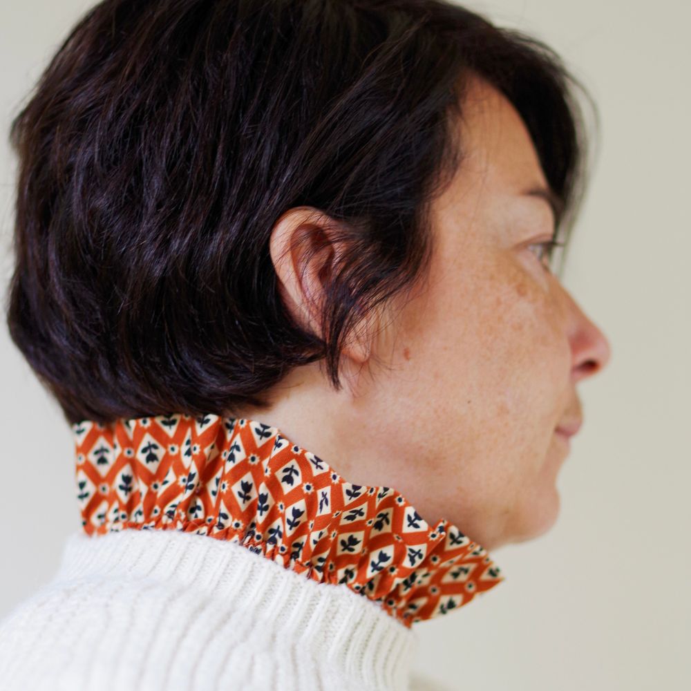 Femme aux cheveux bruns courts, vue de profil, portant un col amovible victorien en tissu roux à motifs géométriques et fleurs noires, qui dépasse de l’encolure d’un pull blanc cassé en maille côtelée.