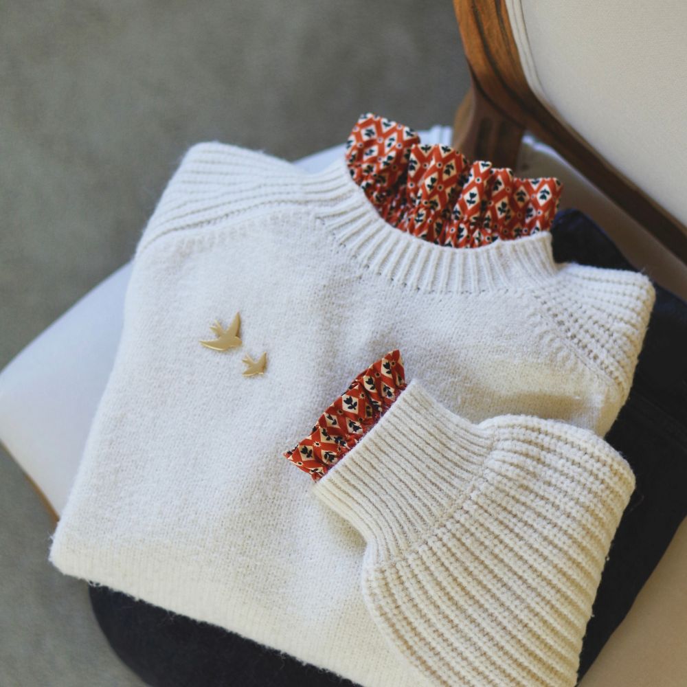 Pull blanc cassé en maille côtelée, plié, avec un col et un poignet amovibles en tissu caramel à motifs géométriques qui dépassent. Deux petites broches hirondelles dorées sont épinglées sur le devant du pull.