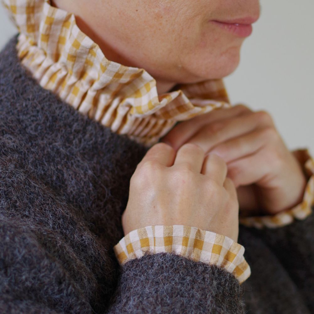 Femme portant un gilet en laine marron glacé, avec un col amovible et des manchettes assortis en vichy mile et nude. Elle porte les mains près du col, dans un geste doux.