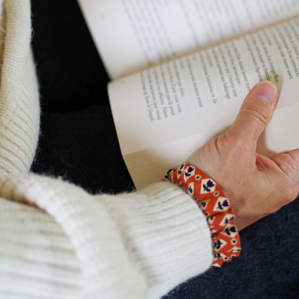 Gros plan sur une main tenant un livre ouvert, avec un poignet agrémenté d'une manchette en tissu roux à motifs géométriques qui dépasse de la manche d’un pull blanc cassé en maille côtelée.