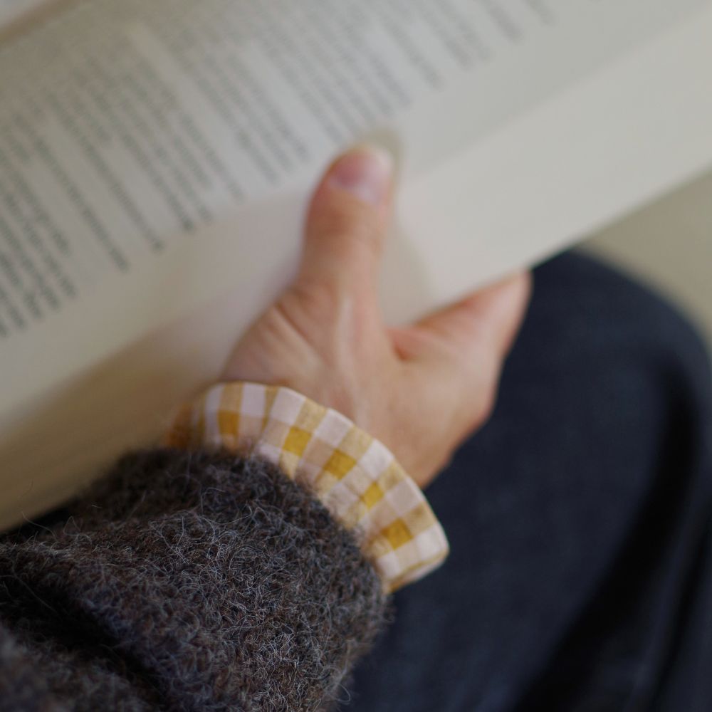 Gros plan sur une main tenant un livre ouvert, avec une manchette amovible en vichy mile et nude qui dépasse de la manche d’un gilet en laine marron glacé. Elle est assise, vêtue d’un jean noir.