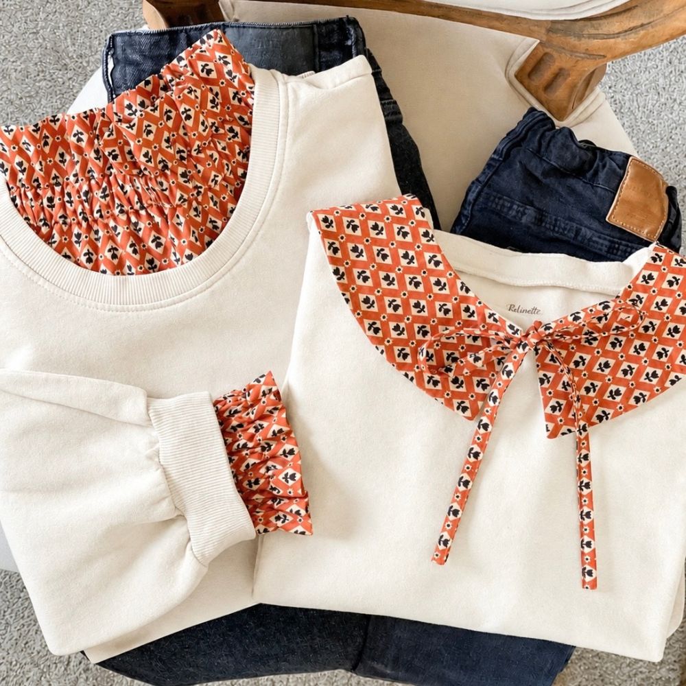 Deux looks matchy-matchy mère-fille composés de sweats écrus pliés côte à côte sur un jean foncé : l’un avec un col victorien et des poignets amovibles en tissu caramel à motifs géométriques blanc et fleurs noires, l’autre avec un col Claudine assorti noué par un ruban.